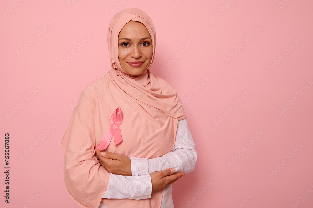 Gorgeous Muslim woman with covered head in hijab, stands on pink background with crossed arms on chest and wearing a pink ribbon, symbol of Breast Cancer Awareness Day. Medical concept, copy space Gorgeous Muslim woman with covered head in hijab, stands on pink background with crossed arms on chest and wearing a pink ribbon, symbol of Breast Cancer Awareness Day. Medical concept, copy space