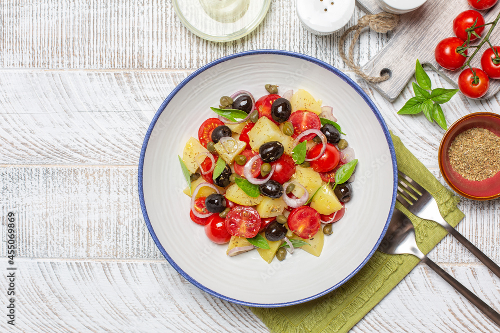 Fresh mediterranean salad.  Made with boiled potatoes, tomatoes, onions, oregano, capers, black olives, basil leaves. White wooden background, top view. Copy space.