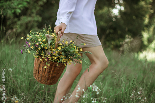 A girl in a white blouse holds a wicker basket with a bouquet of wild flowers. Summer walk in the field. close-up of a woman's legs walking through a field with a basket of flowers