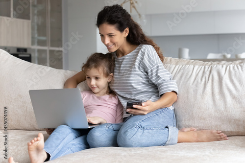 Smiling bonding family young mother and little adorable child daughter spending free weekend leisure time using different devices, resting on comfortable sofa, modern technology addiction concept.