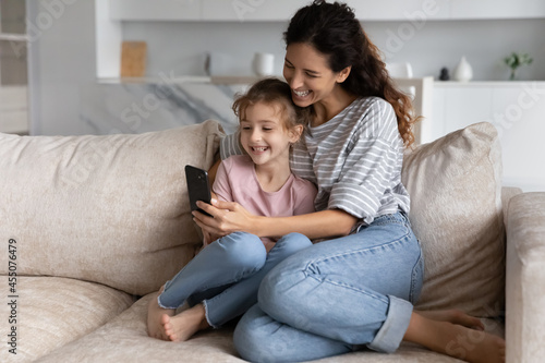 Positive happy young mother and laughing little adorable kid daughter using cellphone applications, recording funny video or posing for selfie photo, resting together on comfortable couch at home.