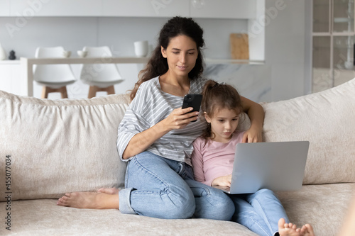 Addicted to modern technology happy young mother and cute little child daughter using different gadgets, spending time online in social networks or playing games, sitting on cozy sofa in living room,
