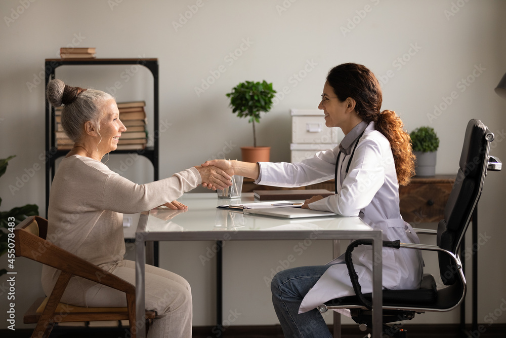 Mature senior patient and young female therapist celebrating successful treatment result with handshake. Old woman visiting doctor, shaking hands, expressing gratitude. Geriatric medic care