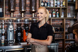 Waitress at the restaurant. The blonde server wears a black t shirt and apron. She has her arms crossed and is looking straight at the camera with a smile. Serving hot and cold drink