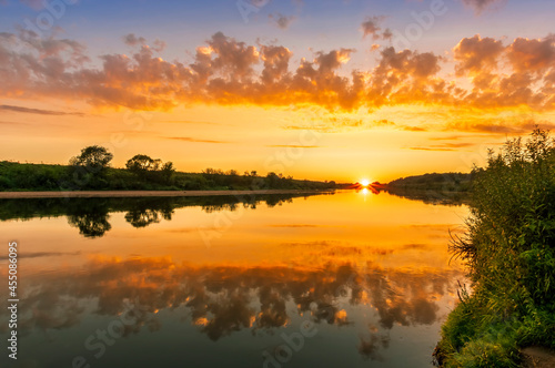Scenic view at beautiful summer river sunset with reflection on water with green bushes, calm water ,deep colorful cloudy sky and glow on horizon on a background, spring evening landscape © Yaroslav