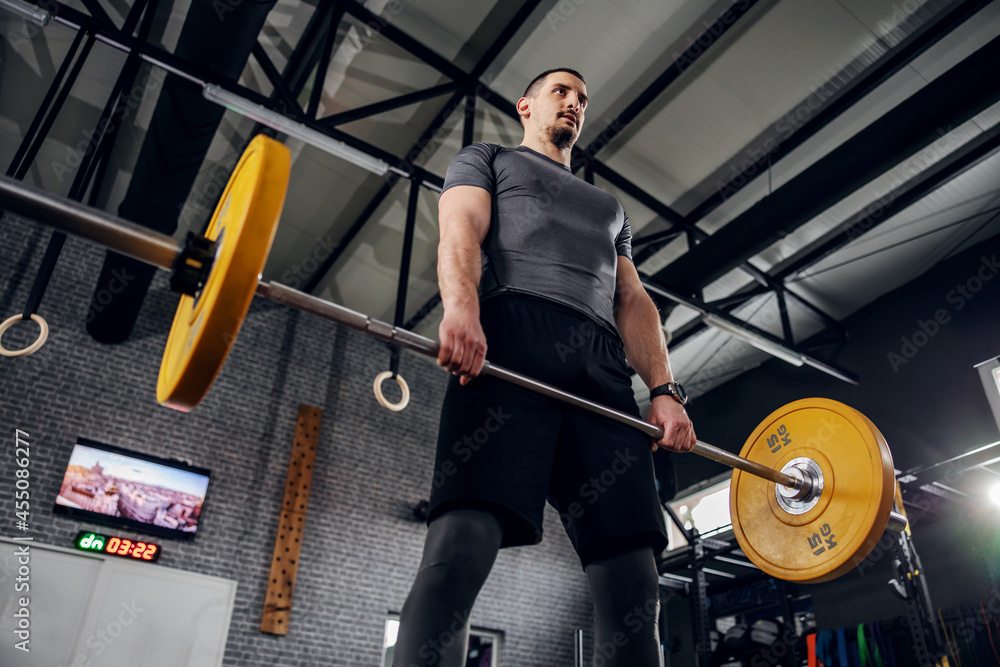Man exercising in a gym with a barbell. Young man in gray sportswear and good physical shape standing in the gym and holding a weighted barbell with both hands and doing exercise. Personal training Man exercising in a gym with a barbell. Young man in gray sportswear and good physical shape standing in the gym and holding a weighted barbell with both hands and doing exercise. Personal training