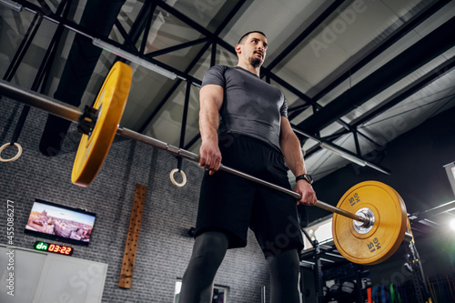 Man exercising in a gym with a barbell. Young man in gray sportswear and good physical shape standing in the gym and holding a weighted barbell with both hands and doing exercise. Personal training