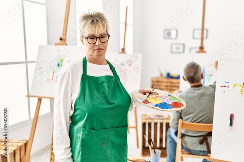 Middle age caucasian woman drawing canvas at art studio thinking attitude and sober expression looking self confident