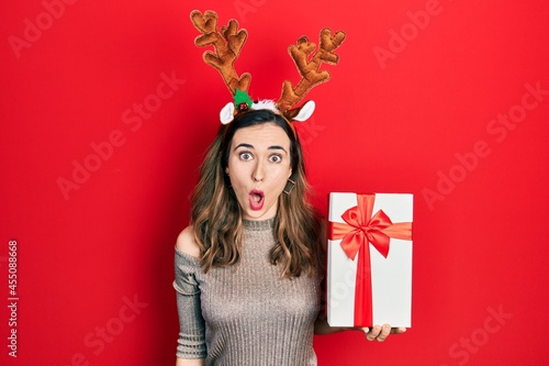 Young hispanic girl wearing deer christmas hat holding gift scared and amazed with open mouth for surprise, disbelief face