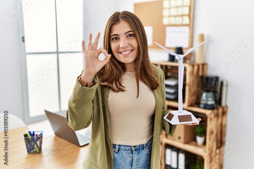 Young brunette woman holding solar windmill for renewable electricity at the office doing ok sign with fingers, smiling friendly gesturing excellent symbol