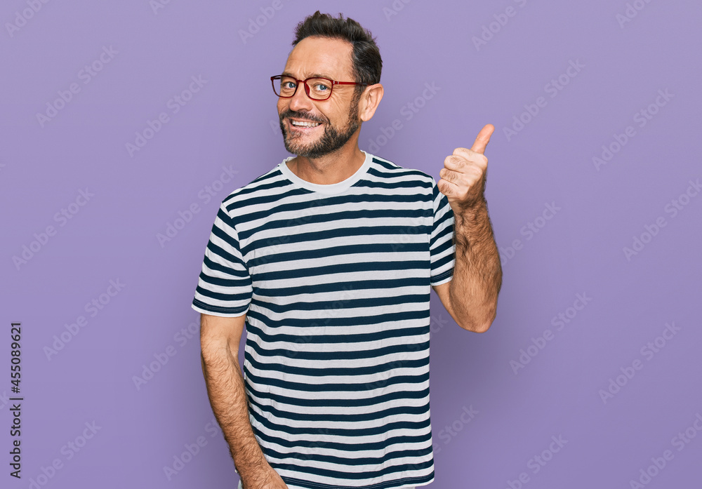 Middle age man wearing casual clothes and glasses smiling with happy face looking and pointing to the side with thumb up. Middle age man wearing casual clothes and glasses smiling with happy face looking and pointing to the side with thumb up.