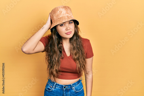 Young hispanic girl wearing casual clothes and hat confuse and wonder about question. uncertain with doubt, thinking with hand on head. pensive concept.