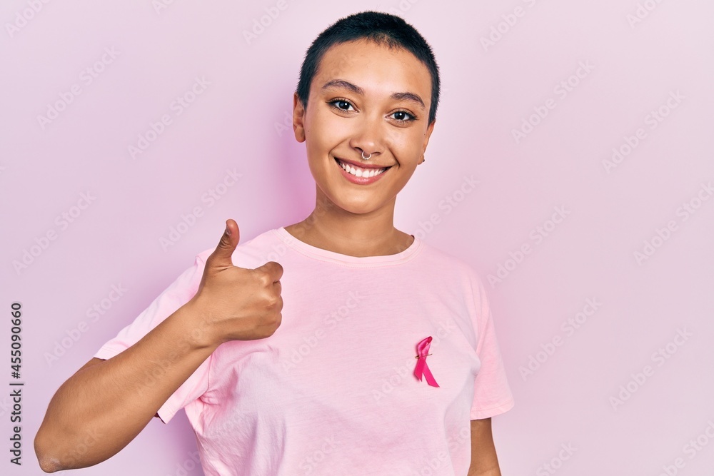 Beautiful hispanic woman with short hair wearing pink cancer ribbon on shirt smiling happy and positive, thumb up doing excellent and approval sign Beautiful hispanic woman with short hair wearing pink cancer ribbon on shirt smiling happy and positive, thumb up doing excellent and approval sign
