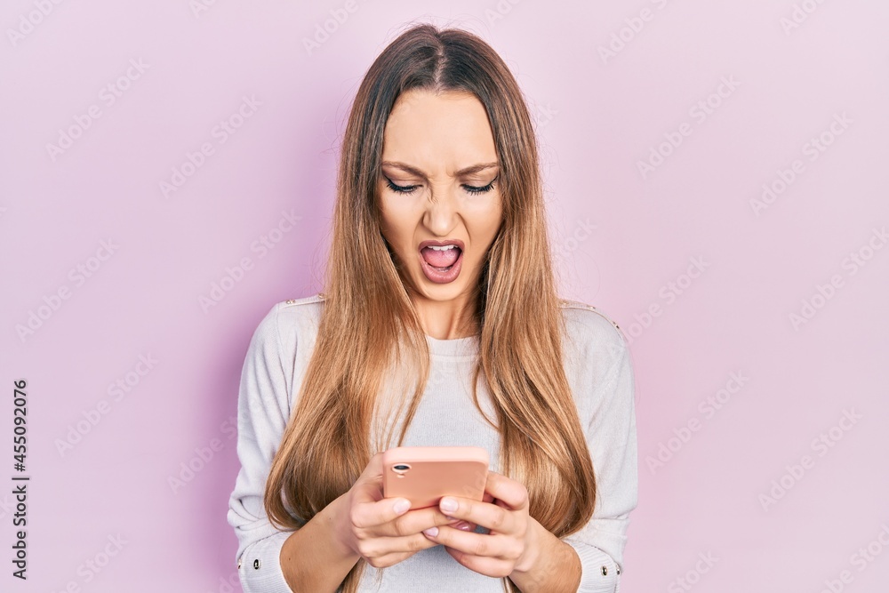 Young blonde girl using smartphone typing message angry and mad screaming frustrated and furious, shouting with anger. rage and aggressive concept.