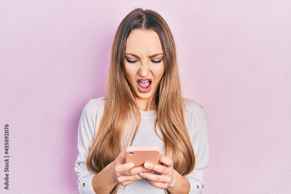 Young blonde girl using smartphone typing message angry and mad screaming frustrated and furious, shouting with anger. rage and aggressive concept. Young blonde girl using smartphone typing message angry and mad screaming frustrated and furious, shouting with anger. rage and aggressive concept.