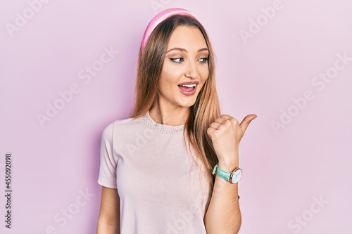 Young blonde girl wearing casual clothes smiling with happy face looking and pointing to the side with thumb up.