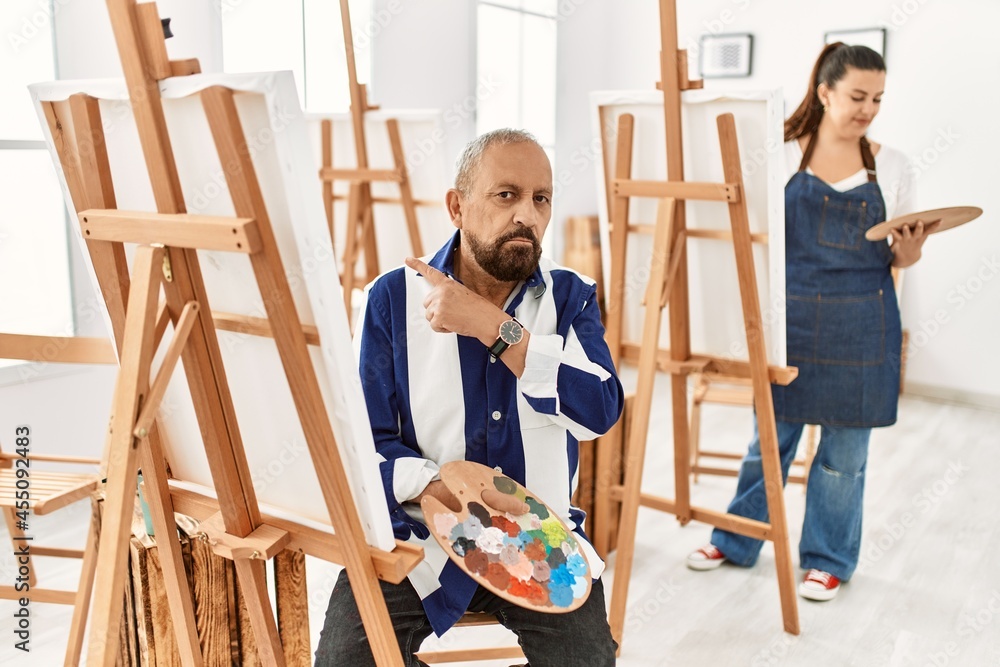 Senior artist man at art studio pointing with hand finger to the side showing advertisement, serious and calm face Senior artist man at art studio pointing with hand finger to the side showing advertisement, serious and calm face