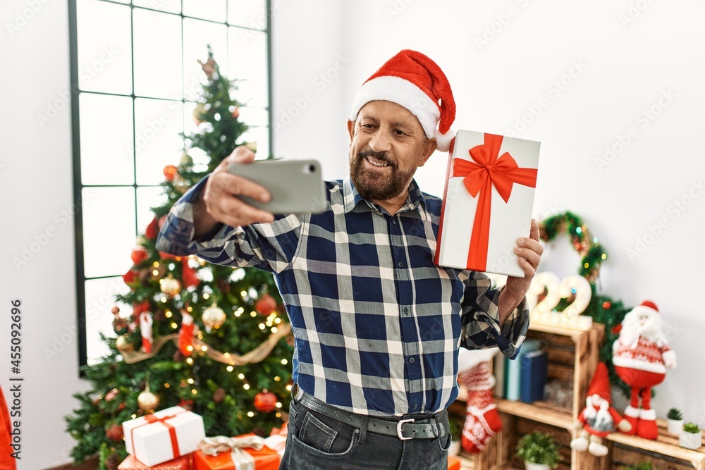 Senior man with beard wearing santa claus hat celebrating christmas at home, standing by christmas tree holding present taking a selfie picture Senior man with beard wearing santa claus hat celebrating christmas at home, standing by christmas tree holding present taking a selfie picture