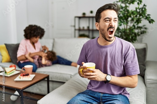 Hispanic father of interracial family drinking a cup coffee angry and mad screaming frustrated and furious, shouting with anger. rage and aggressive concept.