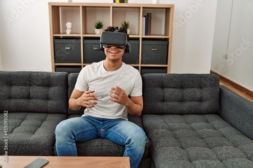 Young hispanic man playing with virtual reality glasses sitting on the sofa at home. photo