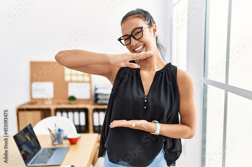 Young hispanic business woman working at the office gesturing with hands showing big and large size sign, measure symbol. smiling looking at the camera. measuring concept.