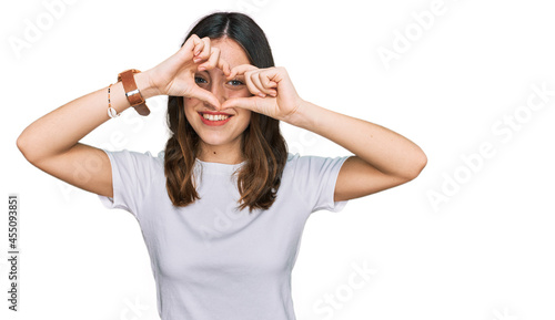 Young beautiful woman wearing casual white t shirt doing heart shape with hand and fingers smiling looking through sign