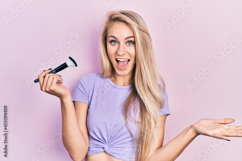 Young caucasian woman holding makeup brush celebrating achievement with happy smile and winner expression with raised hand