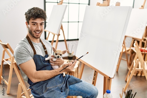 Young hispanic artist man smiling happy holding dog and painting at art studio.