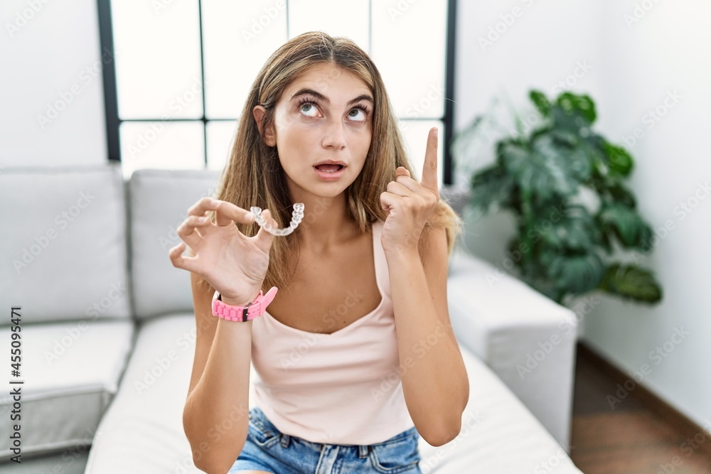 Young blonde woman holding invisible aligner orthodontic amazed and surprised looking up and pointing with fingers and raised arms. Young blonde woman holding invisible aligner orthodontic amazed and surprised looking up and pointing with fingers and raised arms.