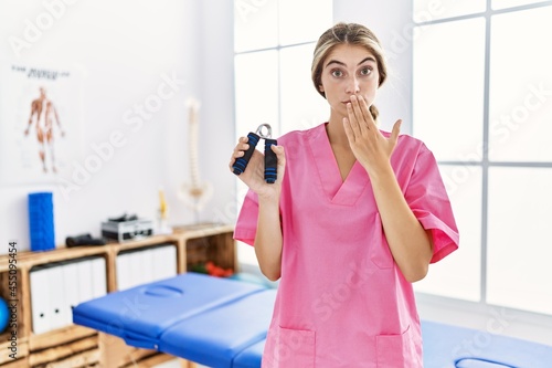 Young blonde woman working at pain recovery clinic holding hand strengthener covering mouth with hand  shocked and afraid for mistake. surprised expression