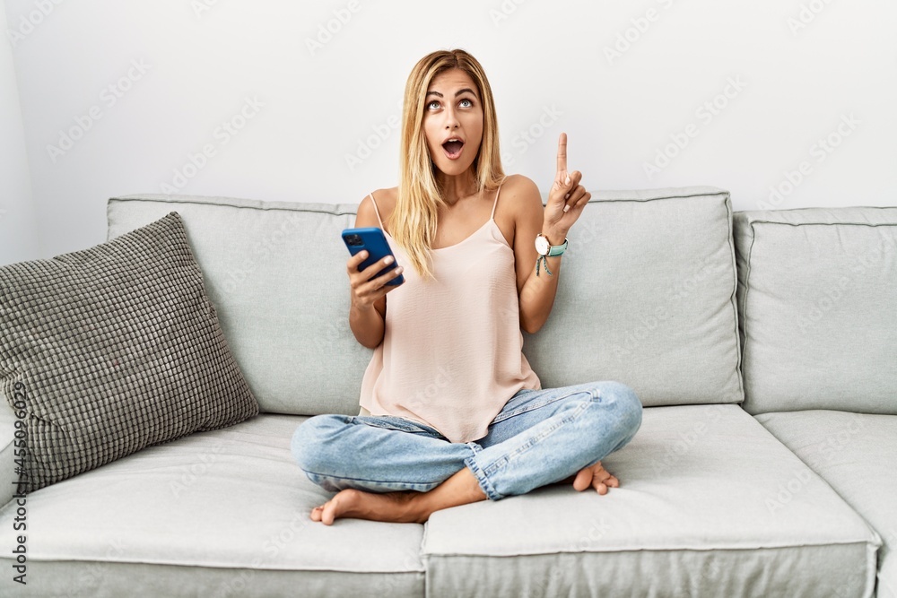 Blonde beautiful young woman sitting on the sofa at home using smartphone amazed and surprised looking up and pointing with fingers and raised arms. Blonde beautiful young woman sitting on the sofa at home using smartphone amazed and surprised looking up and pointing with fingers and raised arms.