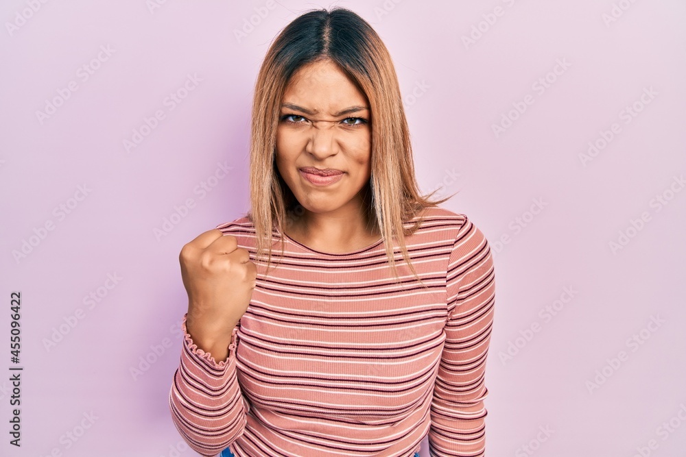 Beautiful hispanic woman wearing casual striped sweater angry and mad raising fist frustrated and furious while shouting with anger. rage and aggressive concept. Beautiful hispanic woman wearing casual striped sweater angry and mad raising fist frustrated and furious while shouting with anger. rage and aggressive concept.