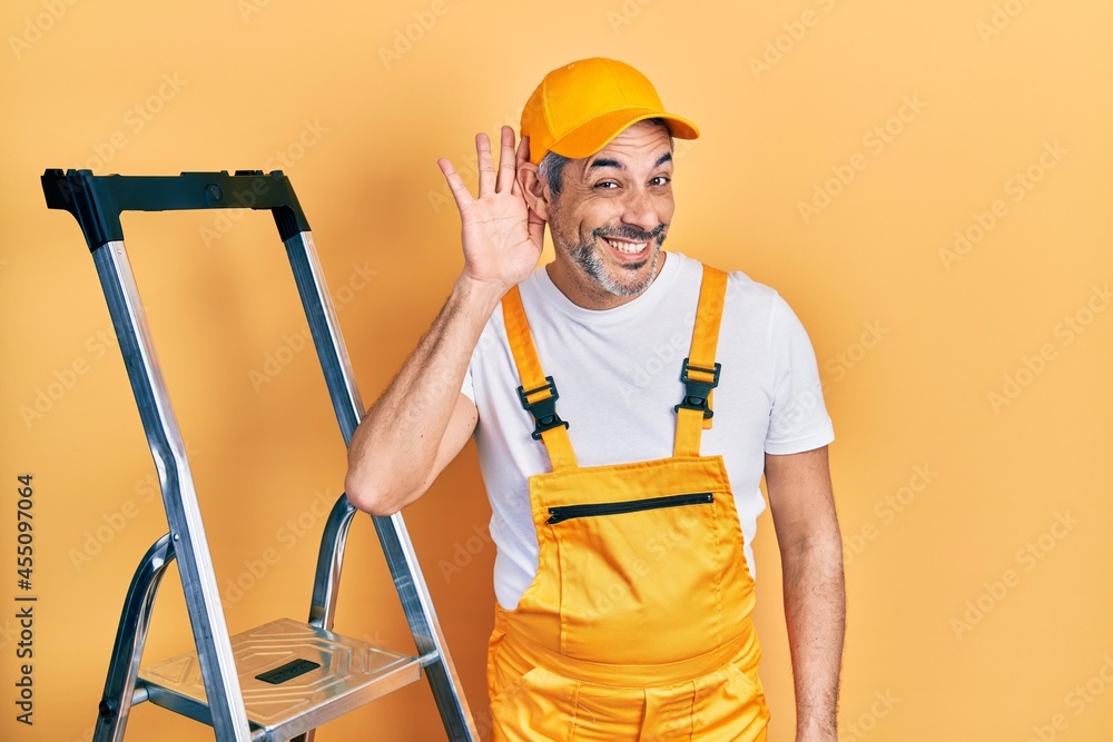 Handsome middle age man with grey hair holding ladder smiling with hand over ear listening an hearing to rumor or gossip. deafness concept. Handsome middle age man with grey hair holding ladder smiling with hand over ear listening an hearing to rumor or gossip. deafness concept.