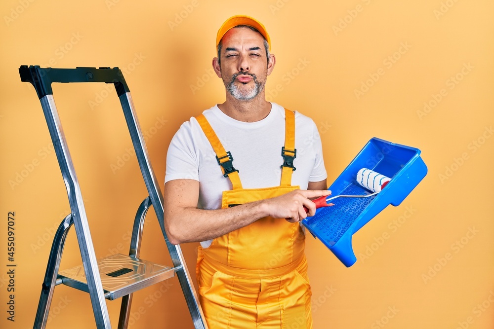 Handsome middle age man with grey hair holding roller painter looking at the camera blowing a kiss being lovely and sexy. love expression. Handsome middle age man with grey hair holding roller painter looking at the camera blowing a kiss being lovely and sexy. love expression.