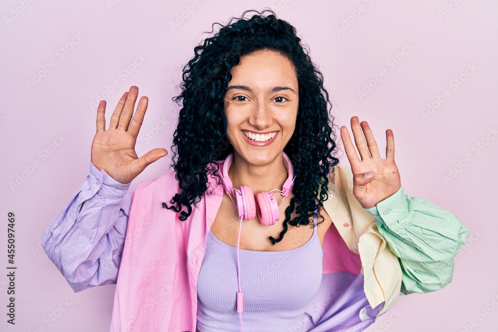 Young hispanic woman with curly hair wearing gym clothes and using headphones showing and pointing up with fingers number nine while smiling confident and happy. Young hispanic woman with curly hair wearing gym clothes and using headphones showing and pointing up with fingers number nine while smiling confident and happy.