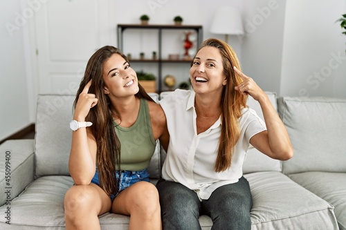 Mother and daughter together sitting on the sofa at home smiling pointing to head with one finger, great idea or thought, good memory