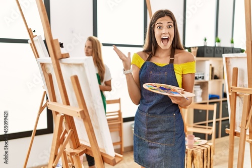 Young hispanic woman at art classroom celebrating victory with happy smile and winner expression with raised hands