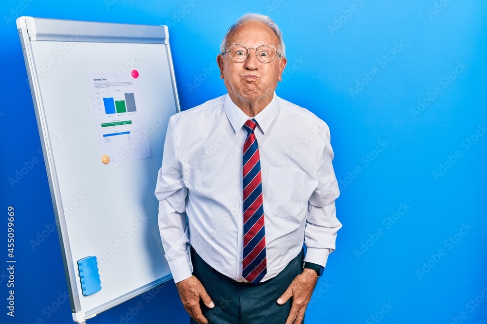 Senior man with grey hair standing by business blackboard puffing cheeks with funny face. mouth inflated with air, crazy expression. Senior man with grey hair standing by business blackboard puffing cheeks with funny face. mouth inflated with air, crazy expression.