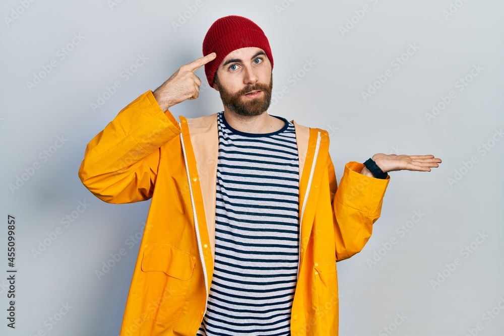 Caucasian man with beard wearing yellow raincoat confused and annoyed with open palm showing copy space and pointing finger to forehead. think about it.