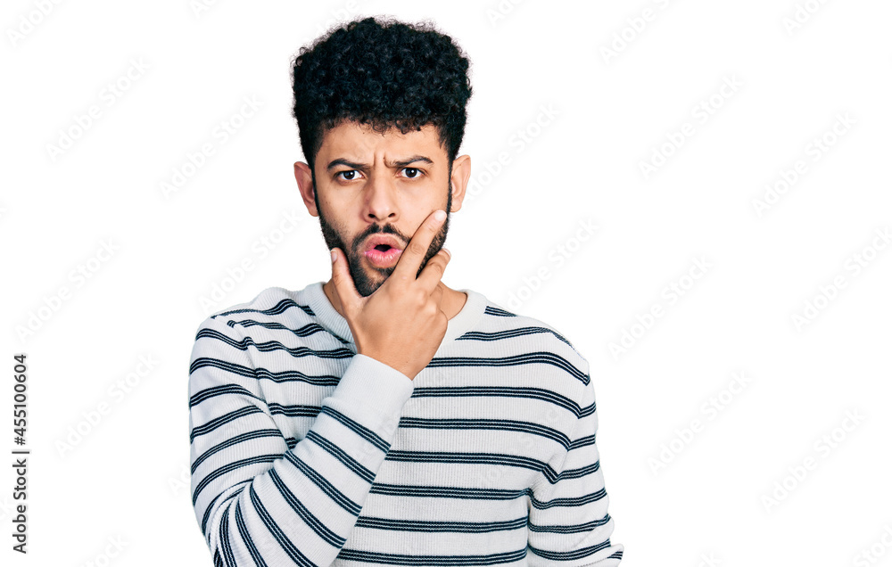 Young arab man with beard wearing casual striped sweater looking fascinated with disbelief, surprise and amazed expression with hands on chin