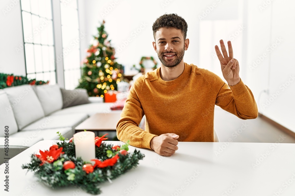 Arab young man sitting on the table by christmas tree showing and pointing up with fingers number three while smiling confident and happy. Arab young man sitting on the table by christmas tree showing and pointing up with fingers number three while smiling confident and happy.