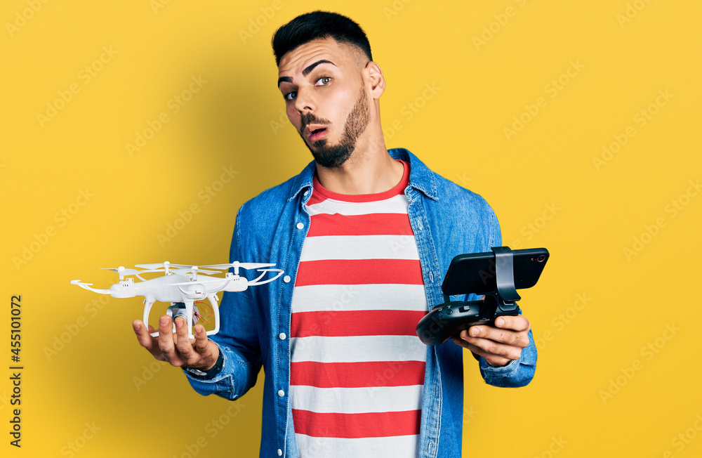 Young hispanic man with beard using drone with remote control in shock face, looking skeptical and sarcastic, surprised with open mouth Young hispanic man with beard using drone with remote control in shock face, looking skeptical and sarcastic, surprised with open mouth