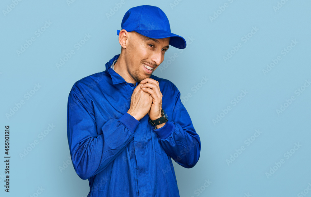 Bald man with beard wearing builder jumpsuit uniform laughing nervous and excited with hands on chin looking to the side