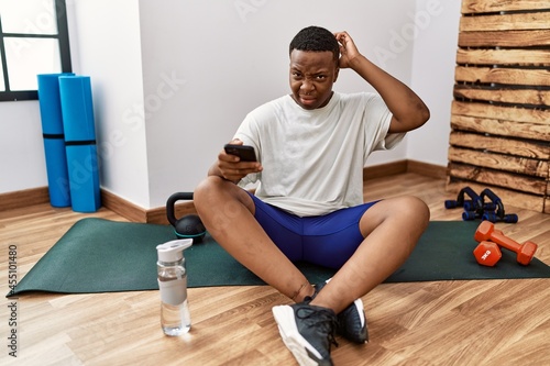 Young african man sitting on training mat at the gym using smartphone confuse and wondering about question. uncertain with doubt, thinking with hand on head. pensive concept.