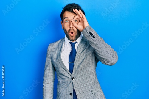 Handsome man with beard wearing business suit and tie doing ok gesture shocked with surprised face, eye looking through fingers. unbelieving expression.