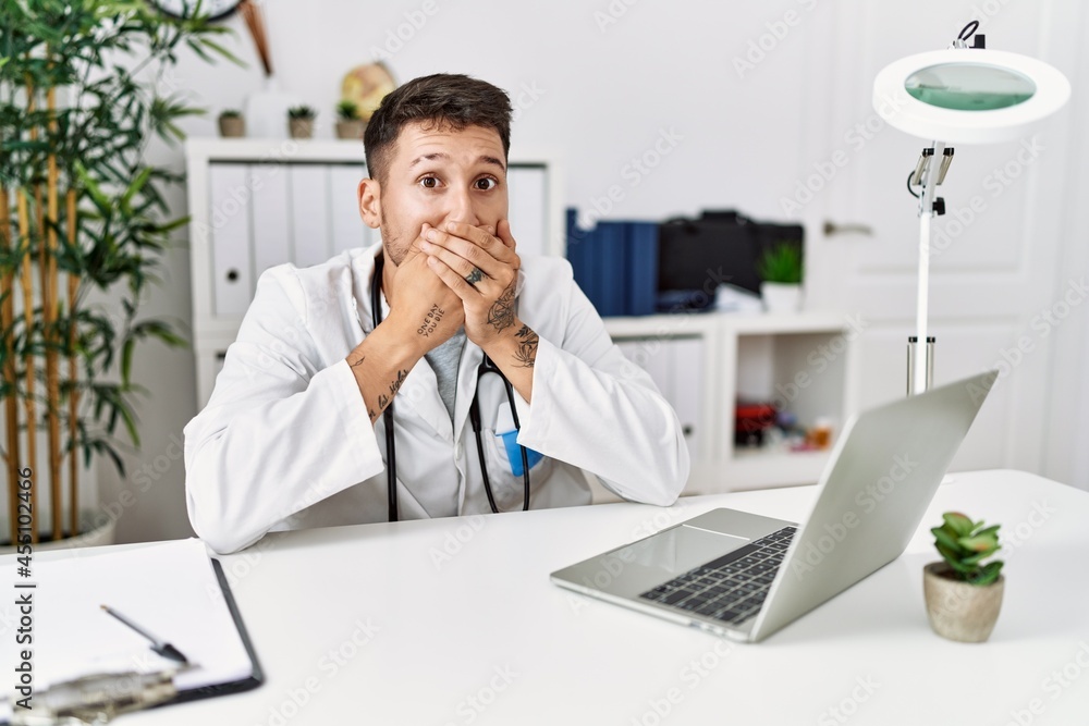 Young doctor working at the clinic using computer laptop shocked covering mouth with hands for mistake. secret concept. Young doctor working at the clinic using computer laptop shocked covering mouth with hands for mistake. secret concept.