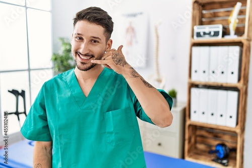 Young physiotherapist man working at pain recovery clinic smiling doing phone gesture with hand and fingers like talking on the telephone. communicating concepts.