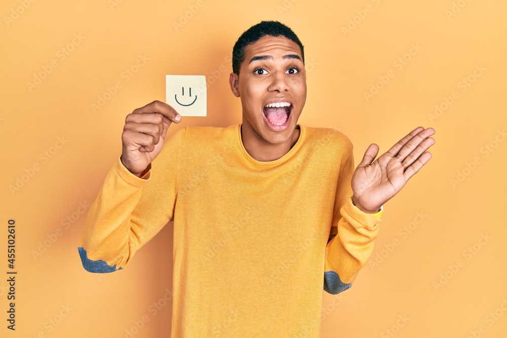 Young african american guy holding smile reminder celebrating achievement with happy smile and winner expression with raised hand Young african american guy holding smile reminder celebrating achievement with happy smile and winner expression with raised hand