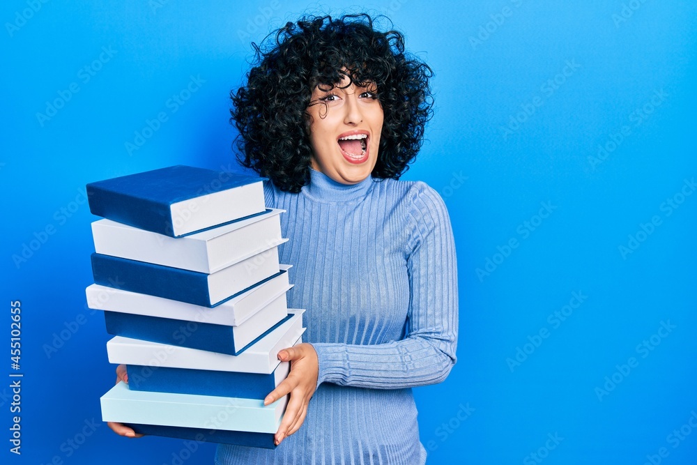 Young middle east woman holding a pile of books celebrating crazy and amazed for success with open eyes screaming excited. Young middle east woman holding a pile of books celebrating crazy and amazed for success with open eyes screaming excited.