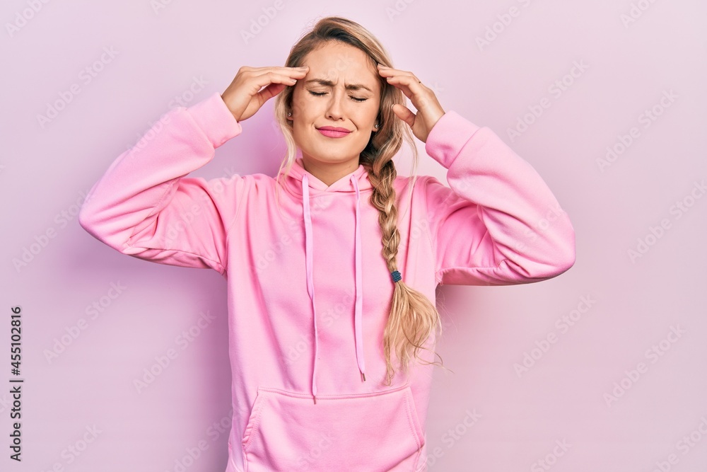 Beautiful young blonde woman wearing pink sweatshirt with hand on head, headache because stress. suffering migraine. Beautiful young blonde woman wearing pink sweatshirt with hand on head, headache because stress. suffering migraine.